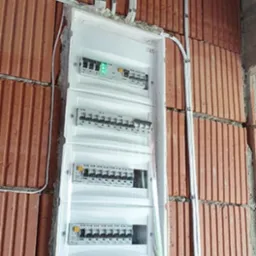 Rozdzielnica elektryczna z zamontowanymi bezpiecznikami na ceglanej ścianie, zabezpieczona folią ochronną, widoczne kable w peszlach.