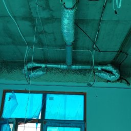 J.K.S company - Montaż instalacji rekuperacji pod sufitem w surowym stanie, widoczne rury wentylacyjne aluminiowe i przewody elektryczne, okno zabezpieczone folią.