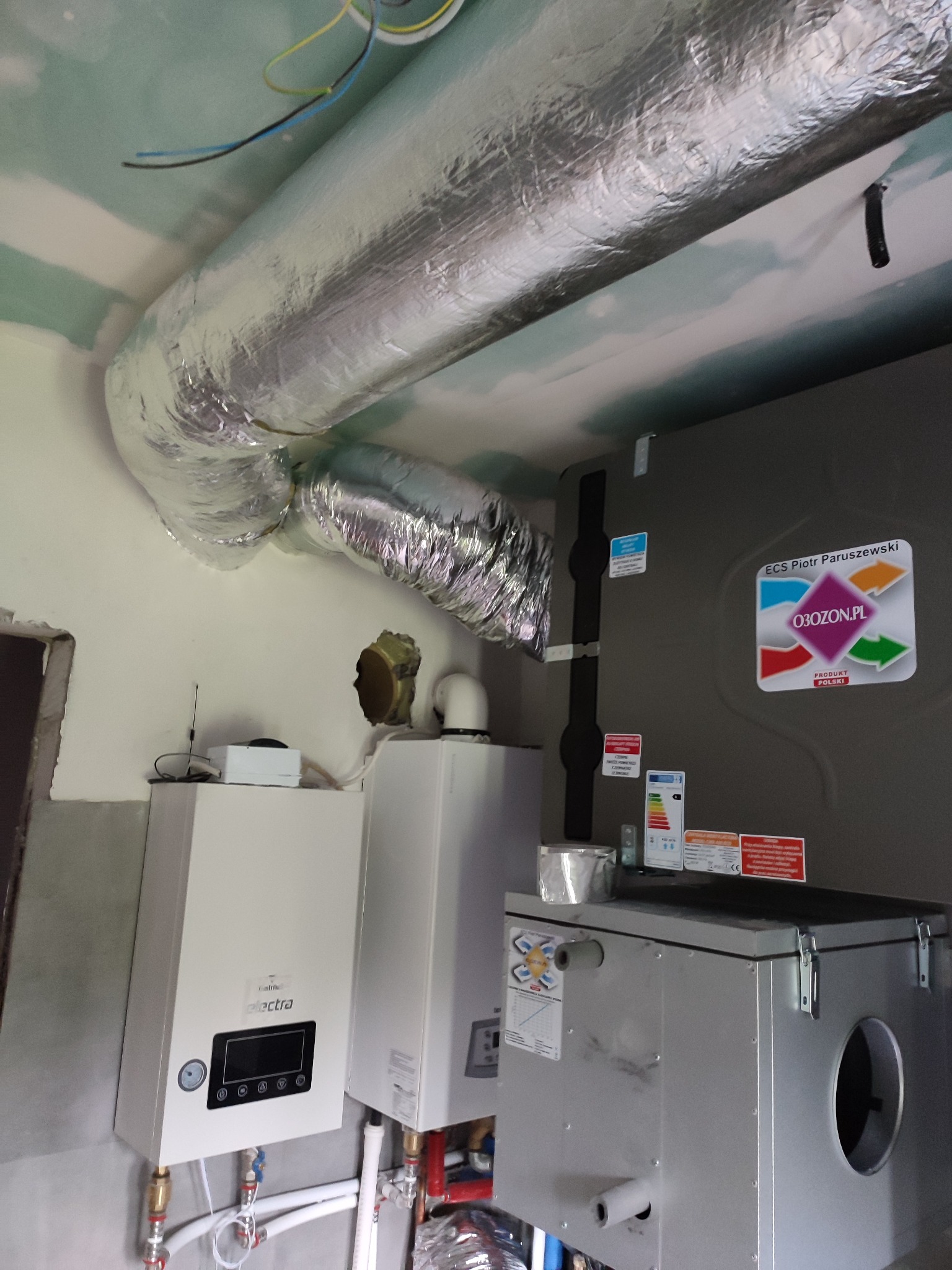 Szczegółowe ujęcie instalacji wentylacyjnej z widocznym rekuperatorem, kanałami wentylacyjnymi w izolacji aluminiowej oraz dwoma białymi urządzeniami grzewczymi, całość w surowym wnętrzu...