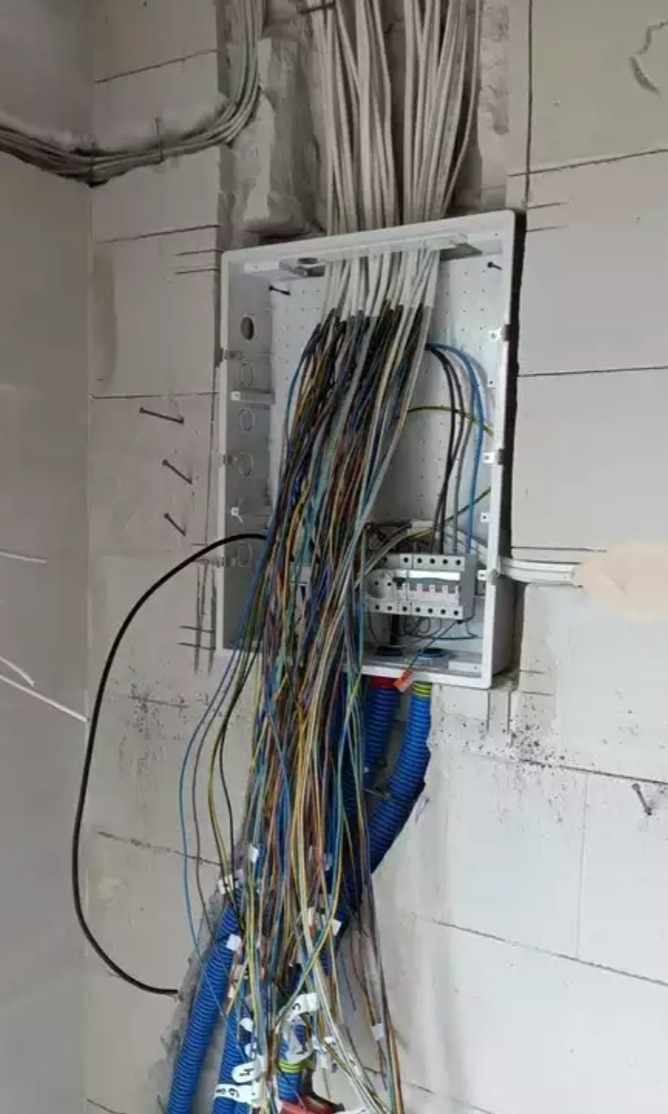 Szafka rozdzielcza z licznymi przewodami elektrycznymi w trakcie montażu w surowym wnętrzu z bloczków betonowych.