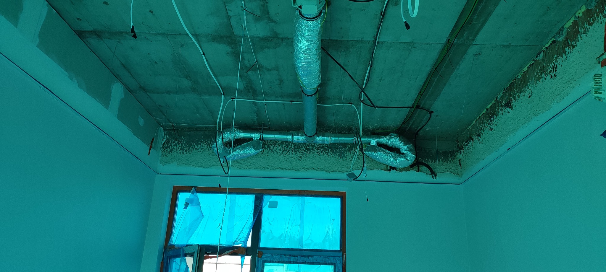 Montaż instalacji rekuperacji pod sufitem w surowym stanie, widoczne rury wentylacyjne aluminiowe i przewody elektryczne, okno zabezpieczone folią.