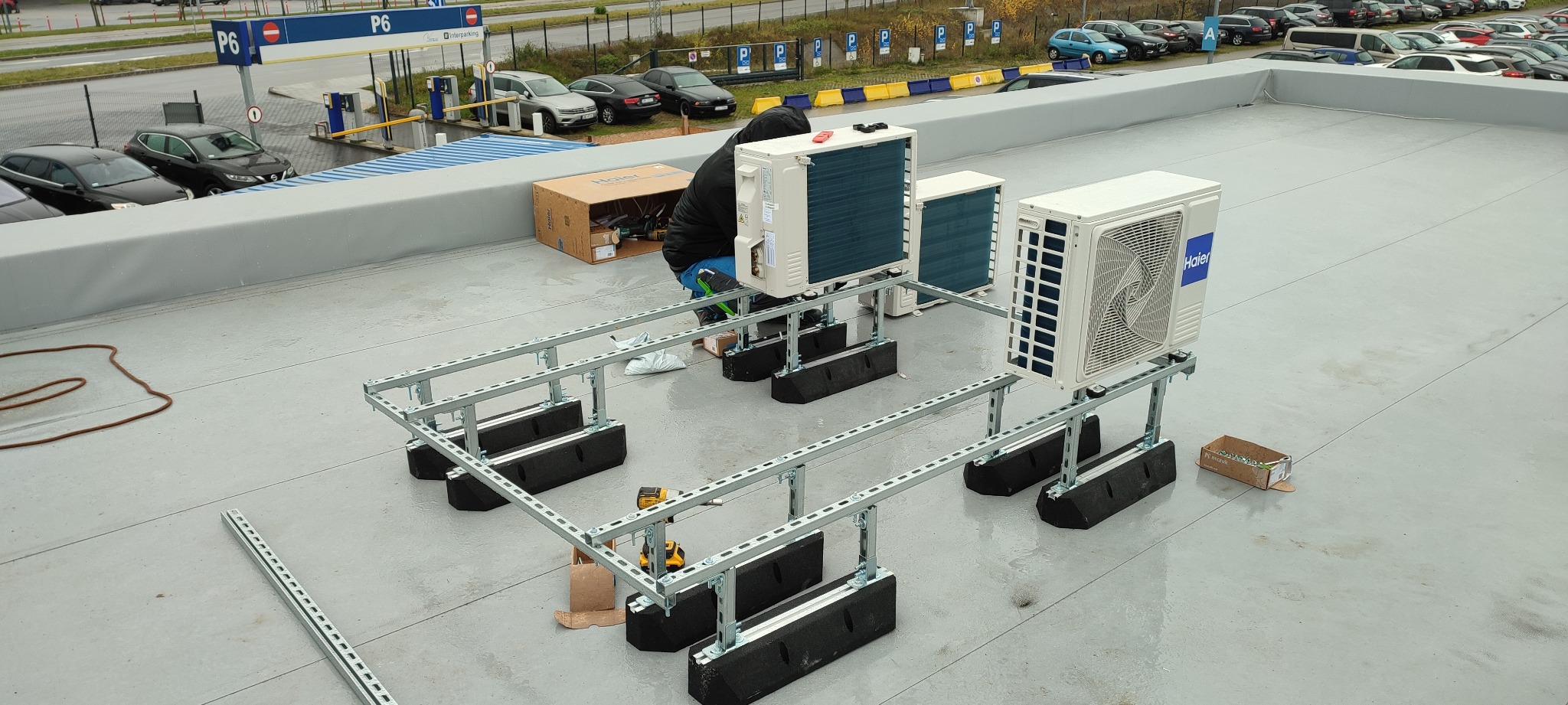 Montaż zewnętrznych jednostek klimatyzacyjnych Haier na dachu budynku. Widoczne metalowe konstrukcje wsporcze z gumowymi podkładkami, narzędzia i pracownik w trakcie instalacji. W tle parking...