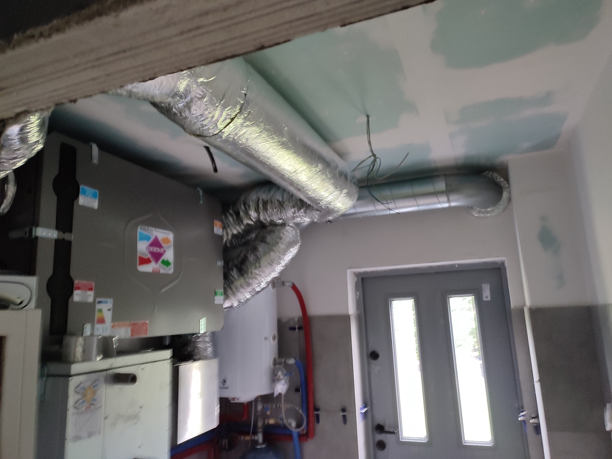 Szare urządzenie z podłączonymi, srebrnymi przewodami wentylacyjnymi w pomieszczeniu z surowym wykończeniem ścian i widoczną instalacją hydrauliczną. Widoczny fragment sufitu z widocznymi śladami...