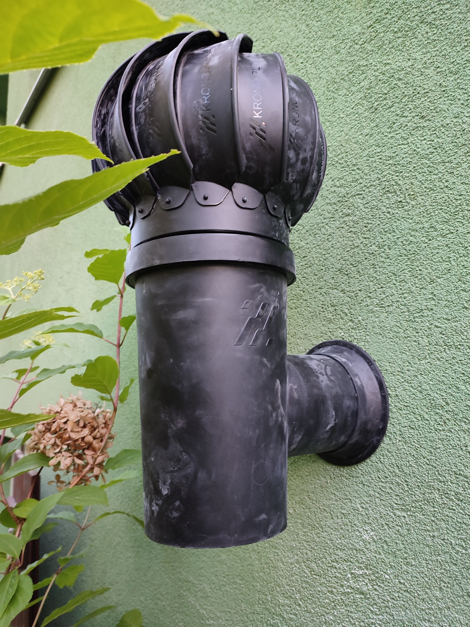 Czarny, obrotowy wywietrznik dachowy marki Krono-Plast zamontowany na elewacji budynku o zielonej, chropowatej strukturze, widoczny z boku, z elementami roślinności w kadrze.