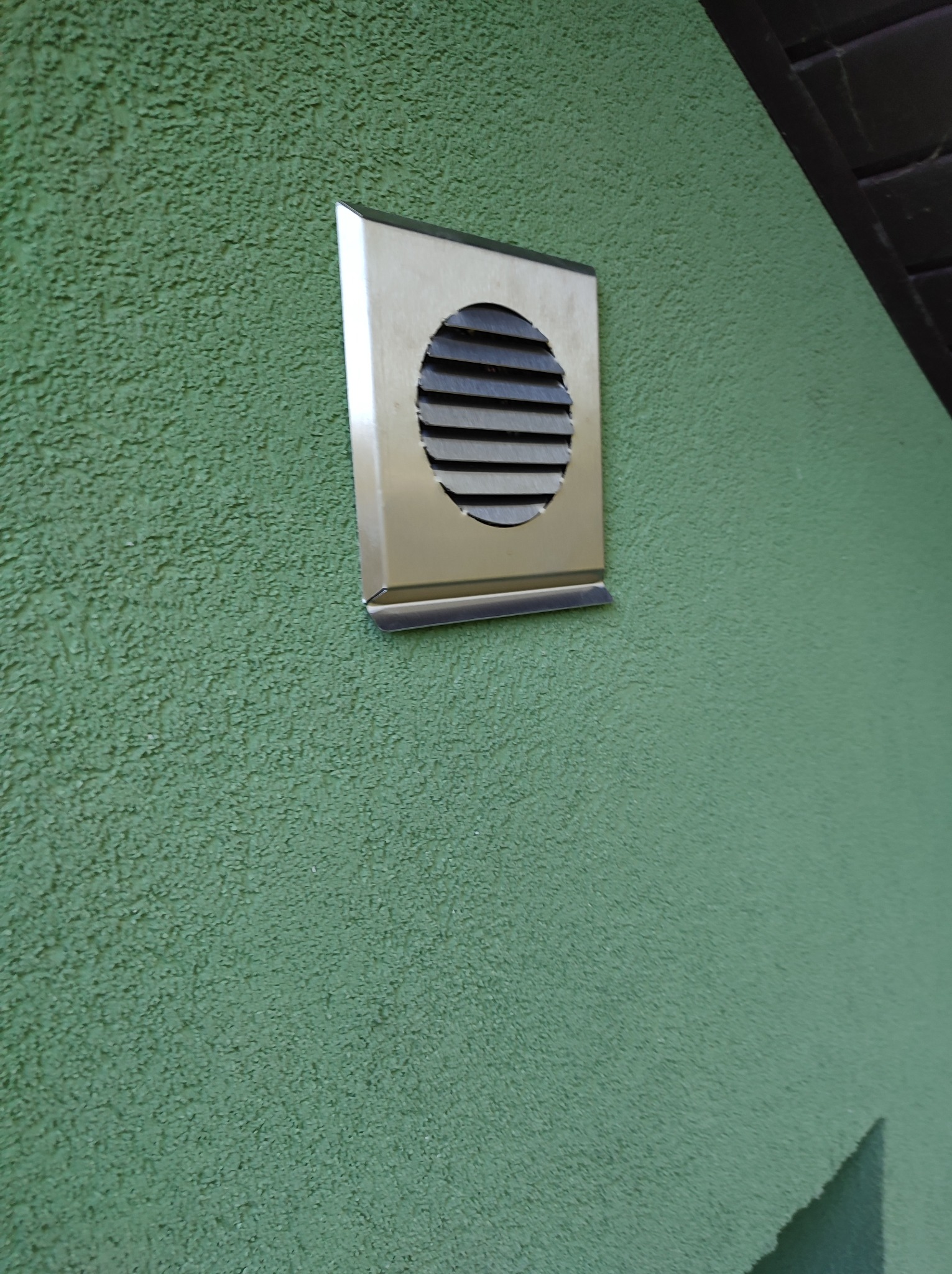 Metalowa kratka wentylacyjna z poziomymi żaluzjami, zamontowana na zielonej, chropowatej ścianie zewnętrznej budynku.