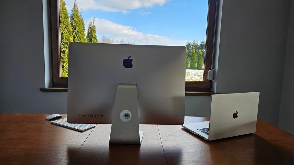 Stanowisko pracy z iMaciem i MacBookiem na biurku z drewna, widok od tyłu, za oknem widoczne zielone drzewa i błękitne niebo z chmurami.