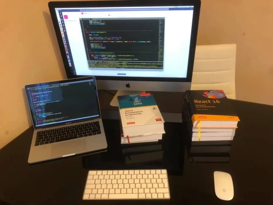 Stanowisko programisty z dwoma ekranami z kodem, książkami do nauki Java i React 16, klawiaturą i myszką na błyszczącym biurku.
