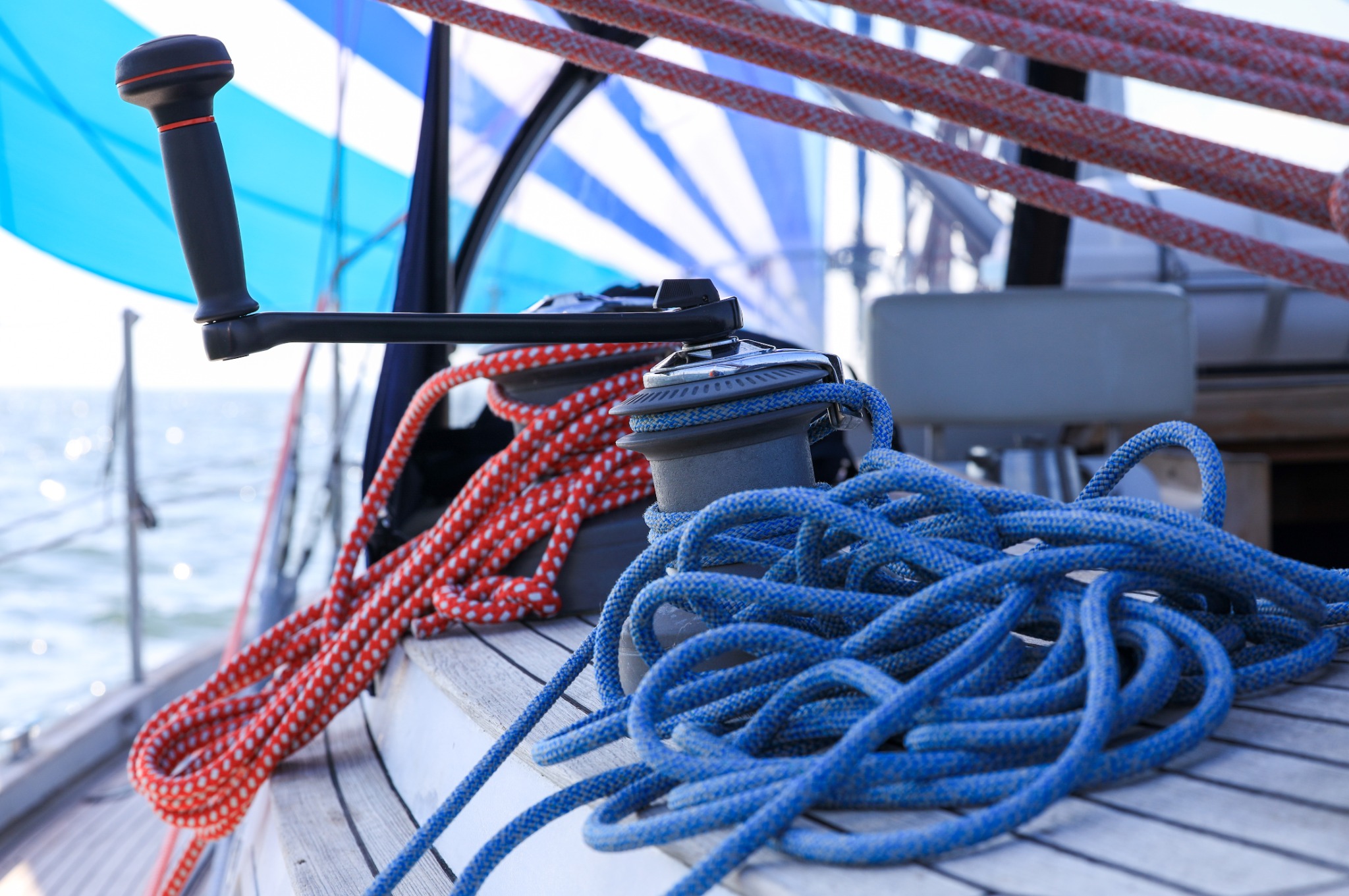 Fragment jachtu z niebiesko-białym żaglem w tle, widoczne liny w kolorach czerwonym i niebieskim, oraz kabestan z korbą.