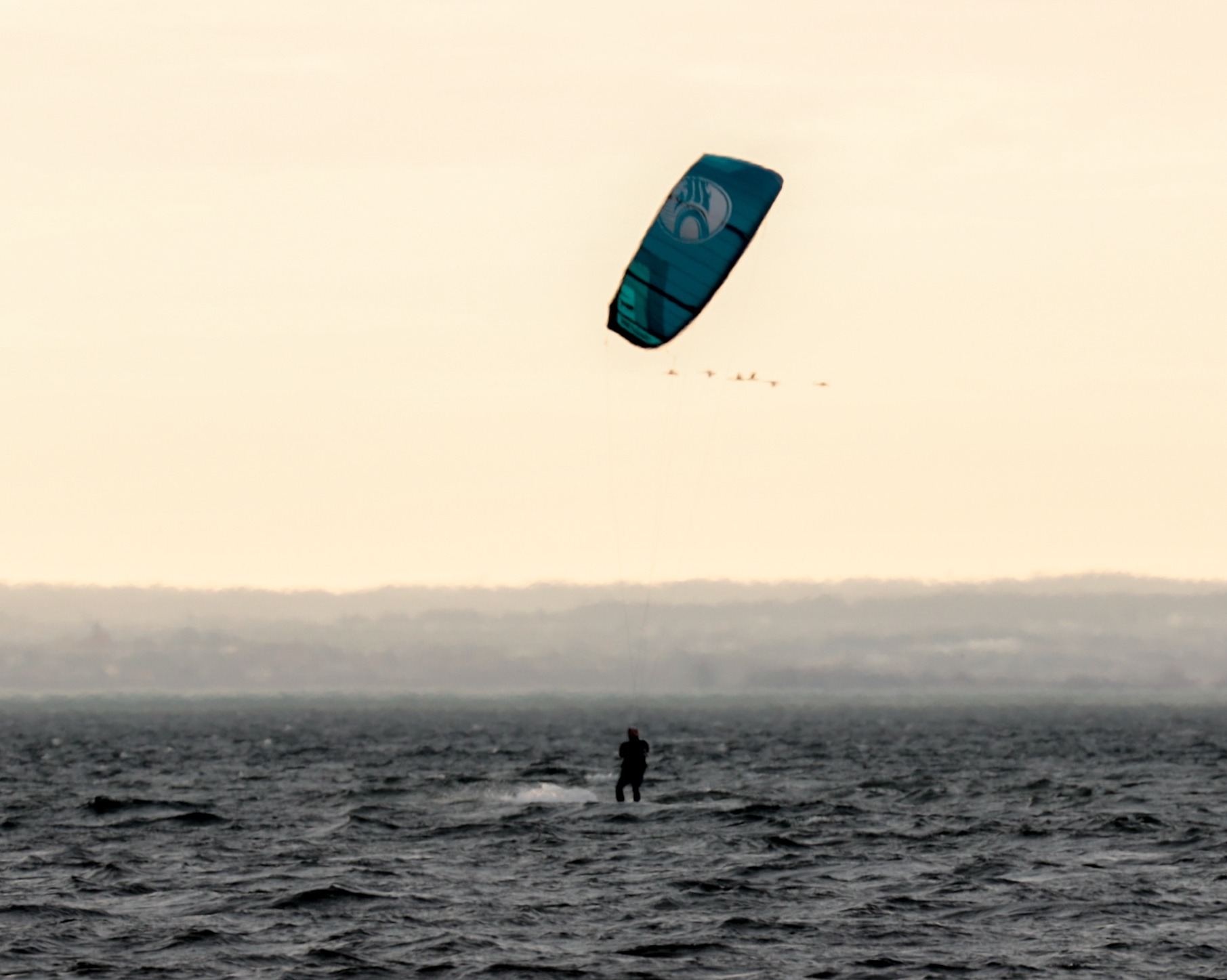 Kite surfer na ciemnym morzu, w tle zarys lądu pod mglistym niebem, nad nim błękitny latawiec i przelatujące ptaki.