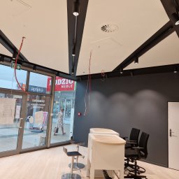 SUPER SIMO MICHAŁ KASPROWSKI - Wnętrze biura w trakcie wykańczania: widoczne niedokończone instalacje elektryczne pod sufitem, surowe ściany pomalowane na szaro, białe biurka i krzesła biurowe, oraz przeszklone wejście...