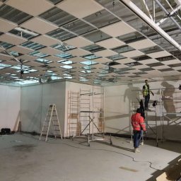 SUPER SIMO MICHAŁ KASPROWSKI - Montaż sufitu podwieszanego w lokalu komercyjnym, pracownicy na rusztowaniach instalują panele, widoczne elementy konstrukcyjne sufitu i oświetlenie.