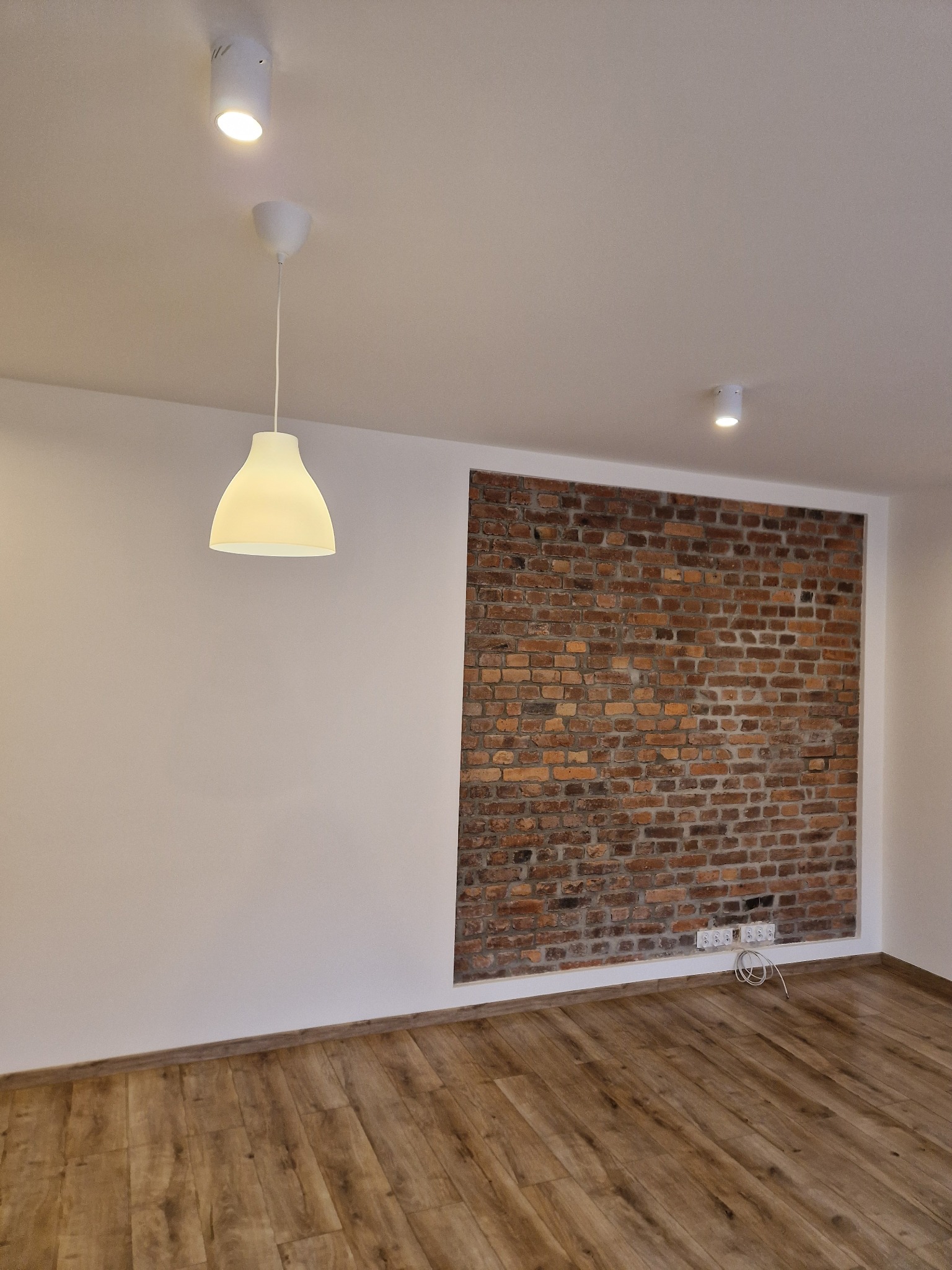 Wnętrze pokoju po odświeżeniu: białe ściany, fragment ściany z cegły, drewniana podłoga, wisząca lampa z białym kloszem i dwa punktowe światła w suficie.