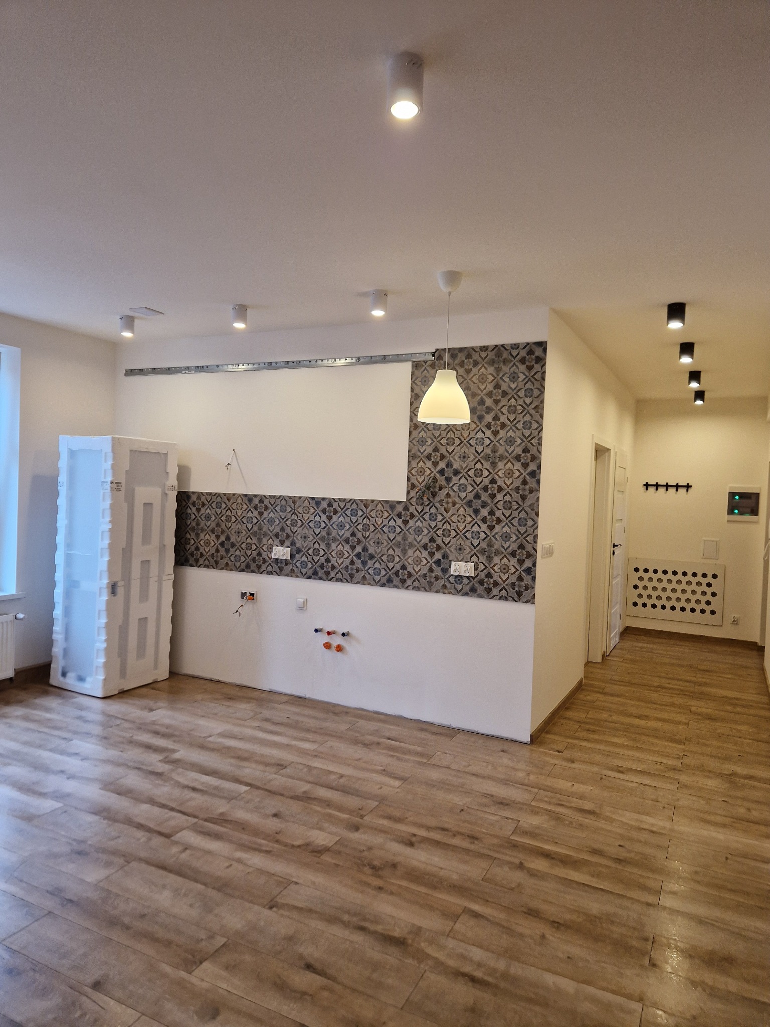 Wnętrze w trakcie prac wykończeniowych: podłoga z paneli imitujących drewno, fragment ściany z ozdobną mozaiką, biała ściana z wystającymi przewodami elektrycznymi i rurkami, zabezpieczona szafa...