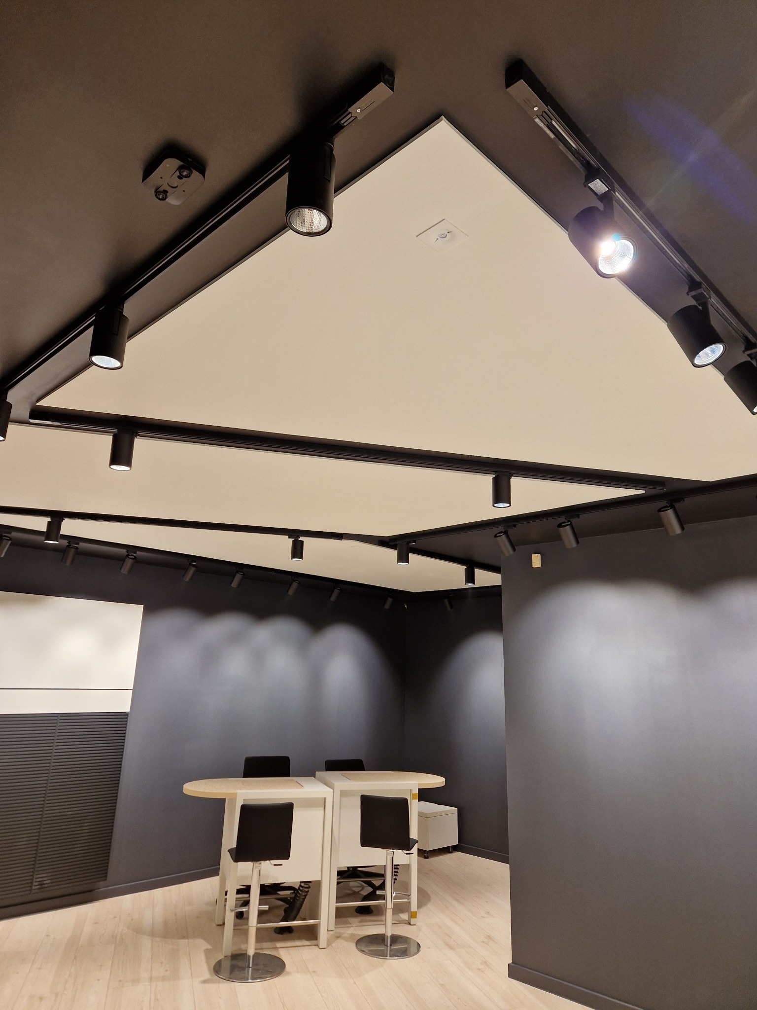 Nowoczesne oświetlenie biurowe: czarne szyny z reflektorami punktowymi na białym suficie kasetonowym w ciemnoszarym biurze z jasną podłogą i wysokimi stołkami barowymi.