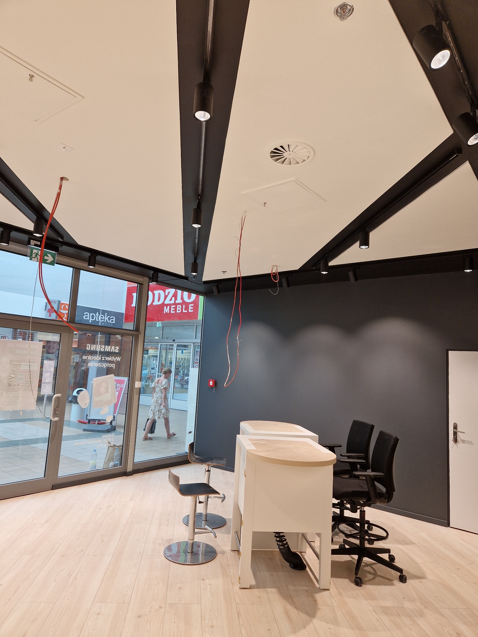 Wnętrze biura w trakcie wykańczania: widoczne niedokończone instalacje elektryczne pod sufitem, surowe ściany pomalowane na szaro, białe biurka i krzesła biurowe, oraz przeszklone wejście...