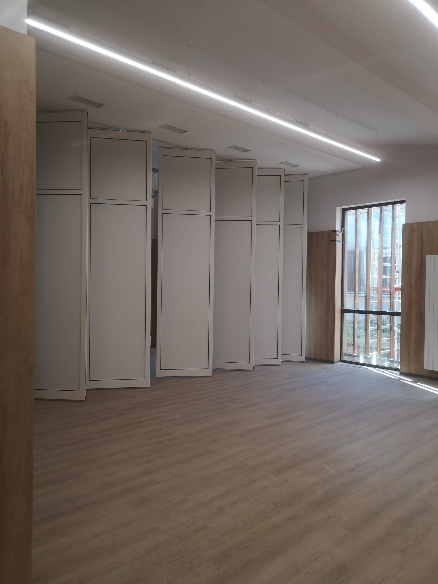 Puste pomieszczenie biurowe z panelami działowymi w trakcie montażu, widoczne drewnopodobne okładziny ścienne i podłoga, oświetlenie liniowe LED.