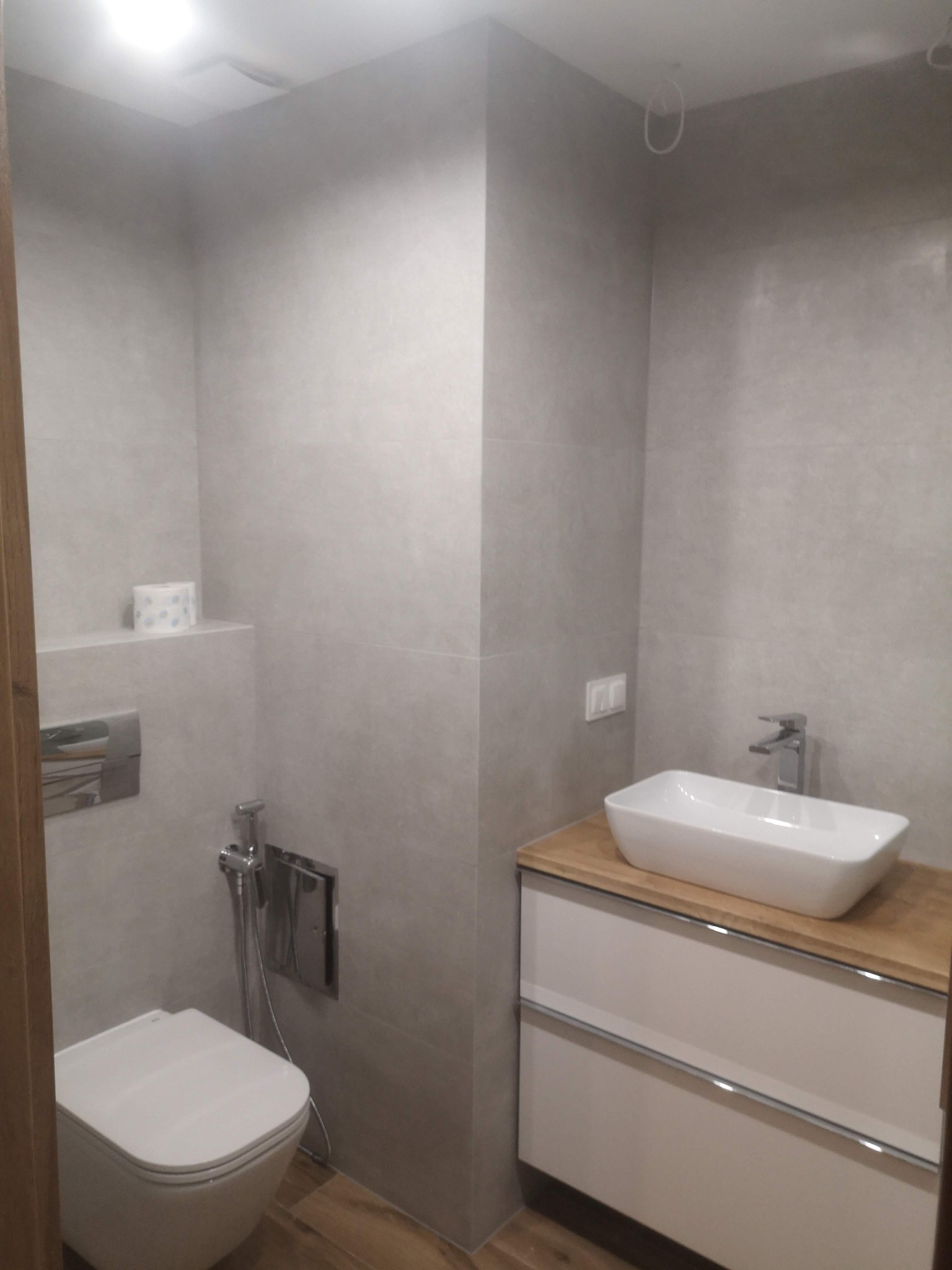 Wykończona łazienka z szarymi płytkami, wiszącą toaletą z bidetką, białą umywalką nablatową na drewnianym blacie i białymi szafkami z chromowanymi uchwytami.