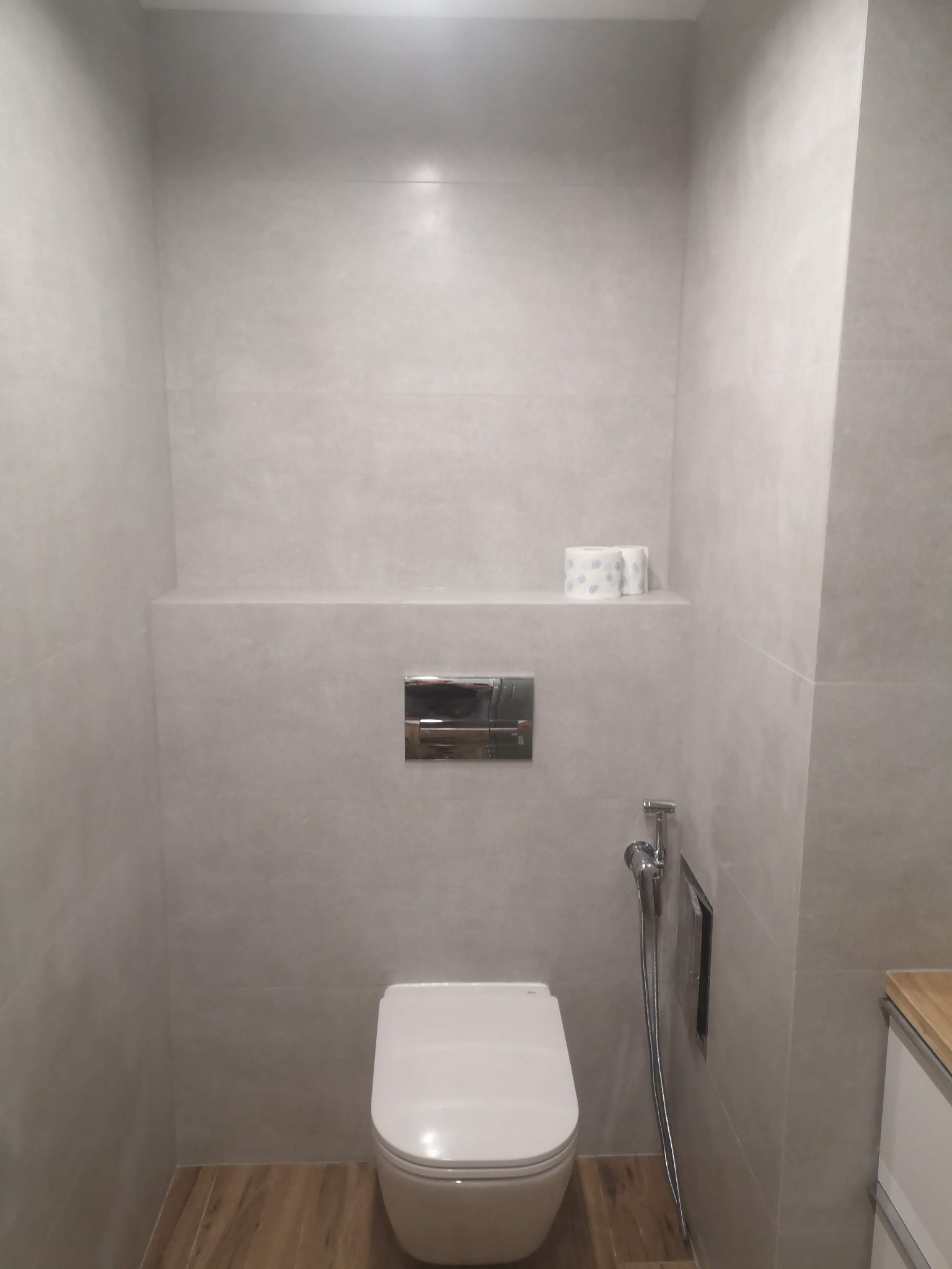 Minimalistyczna toaleta w szaro-beżowej tonacji z podwieszaną muszlą, chromowaną armaturą bidetową i rolkami papieru toaletowego na półce wbudowanej w ścianę wykończoną płytkami.