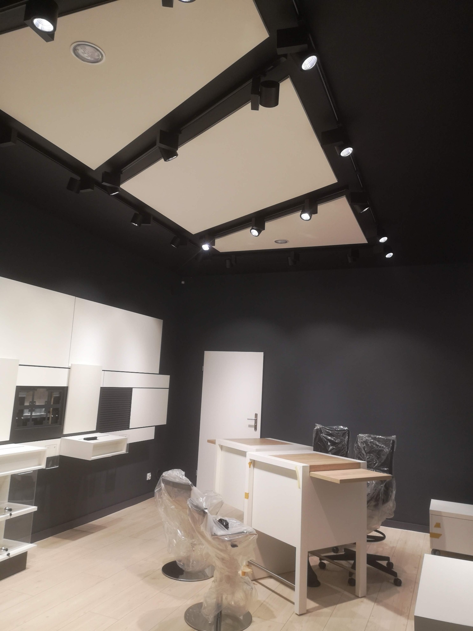 Nowoczesne biuro z czarnymi ścianami i białymi meblami, oświetlenie punktowe na suficie, krzesła biurowe w folii ochronnej, jasna podłoga.