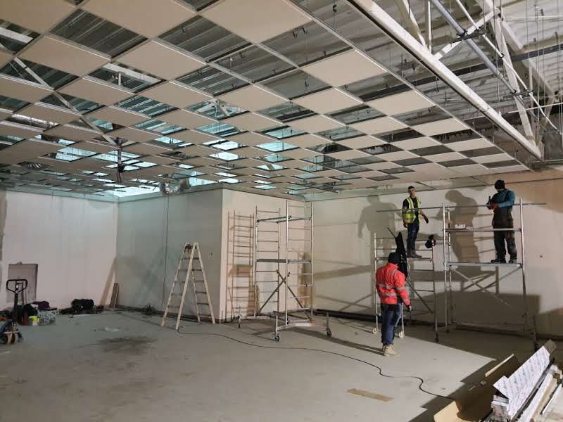 Montaż sufitu podwieszanego w lokalu komercyjnym, pracownicy na rusztowaniach instalują panele, widoczne elementy konstrukcyjne sufitu i oświetlenie.