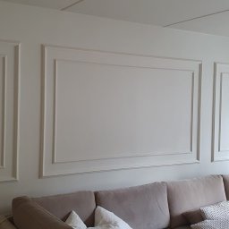 Smart Hangar - Ściana w salonie z dekoracyjnymi, prostokątnymi panelami ściennymi w stylu klasycystycznym, pomalowana na jasny odcień bieli, z widoczną częścią sofy w kolorze beżowym i białymi zasłonami w tle.