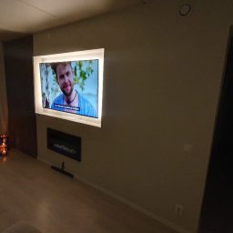 Smart Hangar - Telewizor z podświetleniem LED zamontowany na ścianie nad elektrycznym kominkiem, widoczne dekoracyjne świece i fragment okna z zasłoną.