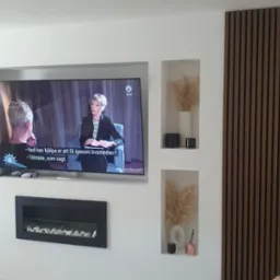 Telewizor wbudowany w ścianę z wnękami dekoracyjnymi, obok pionowy panel z drewnianych listew. W dolnej części czarny, prostokątny kominek elektryczny z białymi kamieniami.