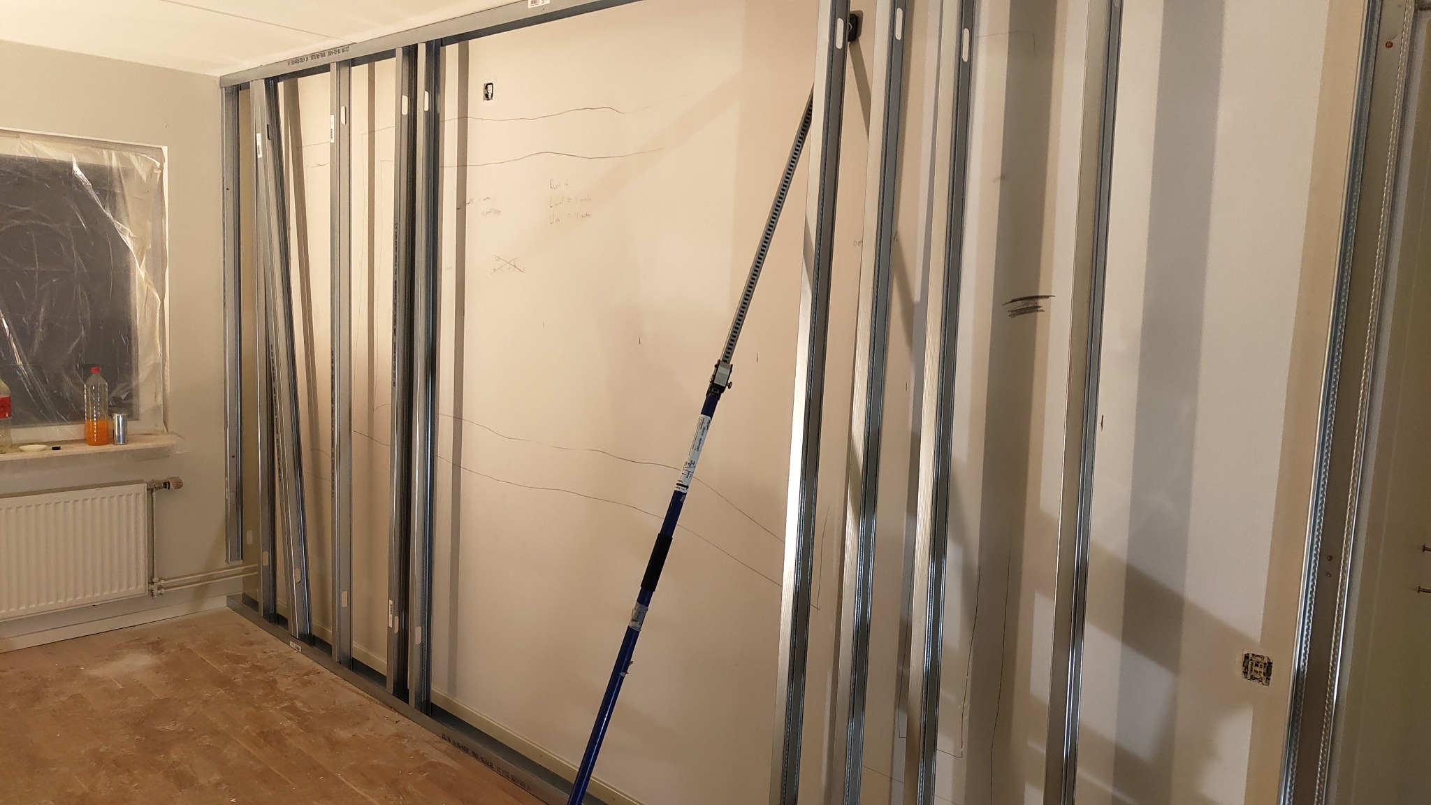 Metalowy szkielet ściany działowej z profili, wsparty rozpórką teleskopową, widoczne oznaczenia na ścianie, okno zabezpieczone folią malarską, grzejnik pod oknem, surowa podłoga.