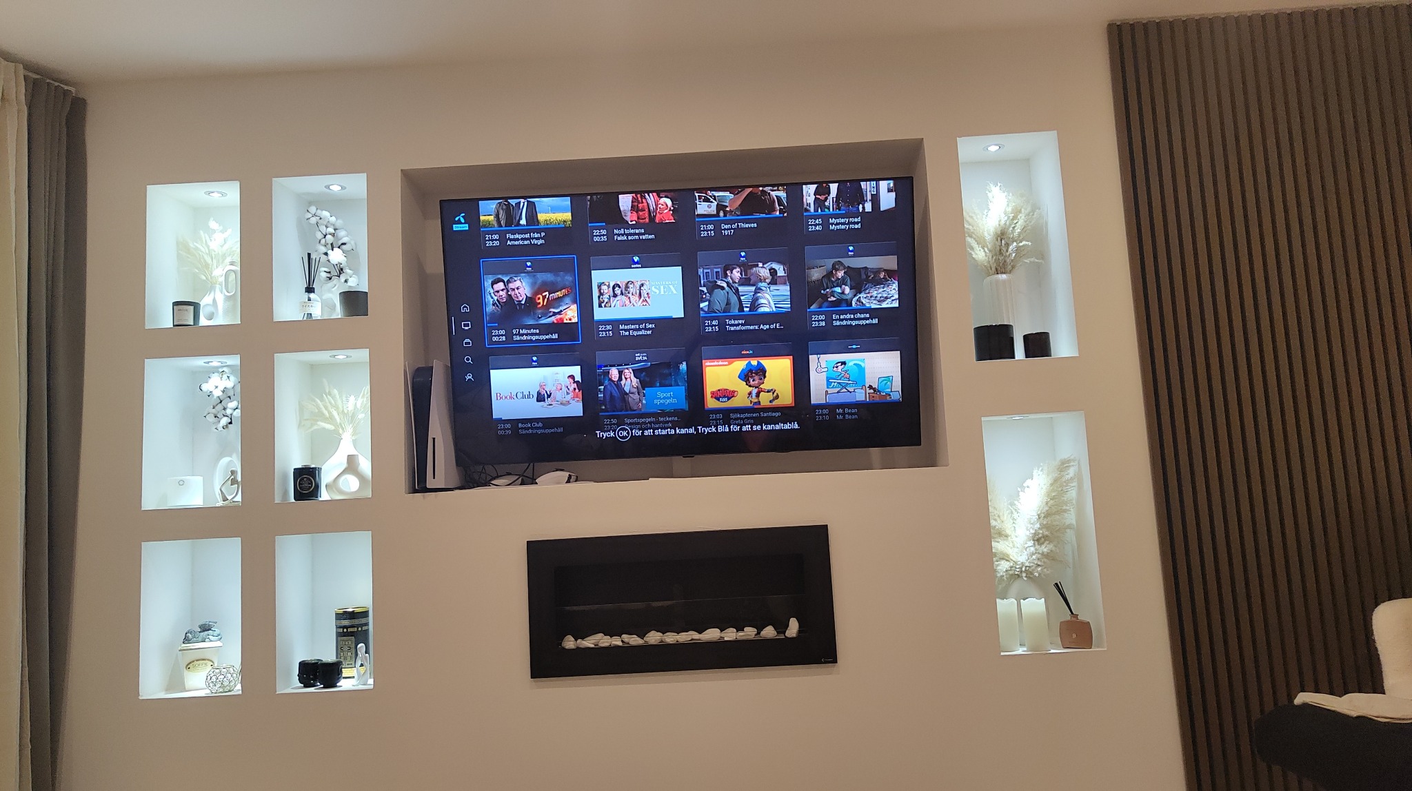 Biała ściana z wnękami oświetlonymi punktowo, w których znajdują się dekoracje: wazony z pampasową trawą, świece i inne ozdoby. Centralnie umieszczony telewizor nad czarnym, prostokątnym kominkiem...