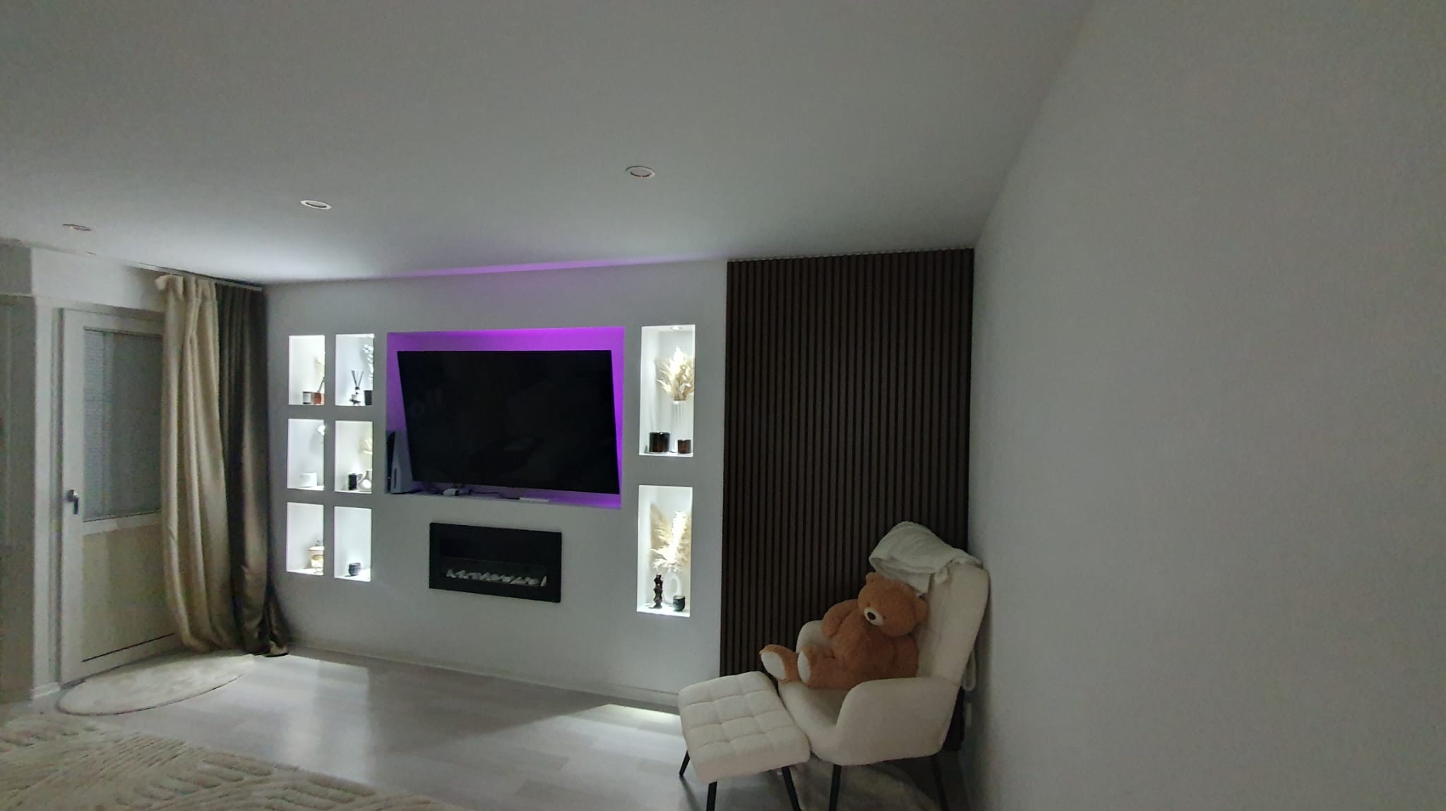 Nowoczesny salon z podświetlonym telewizorem, wnękami dekoracyjnymi, panelem ściennym z drewna i fotelem z pluszowym misiem.