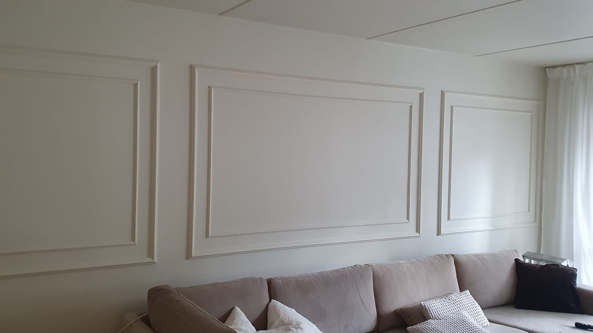 Ściana w salonie z dekoracyjnymi, prostokątnymi panelami ściennymi w stylu klasycystycznym, pomalowana na jasny odcień bieli, z widoczną częścią sofy w kolorze beżowym i białymi zasłonami w tle.