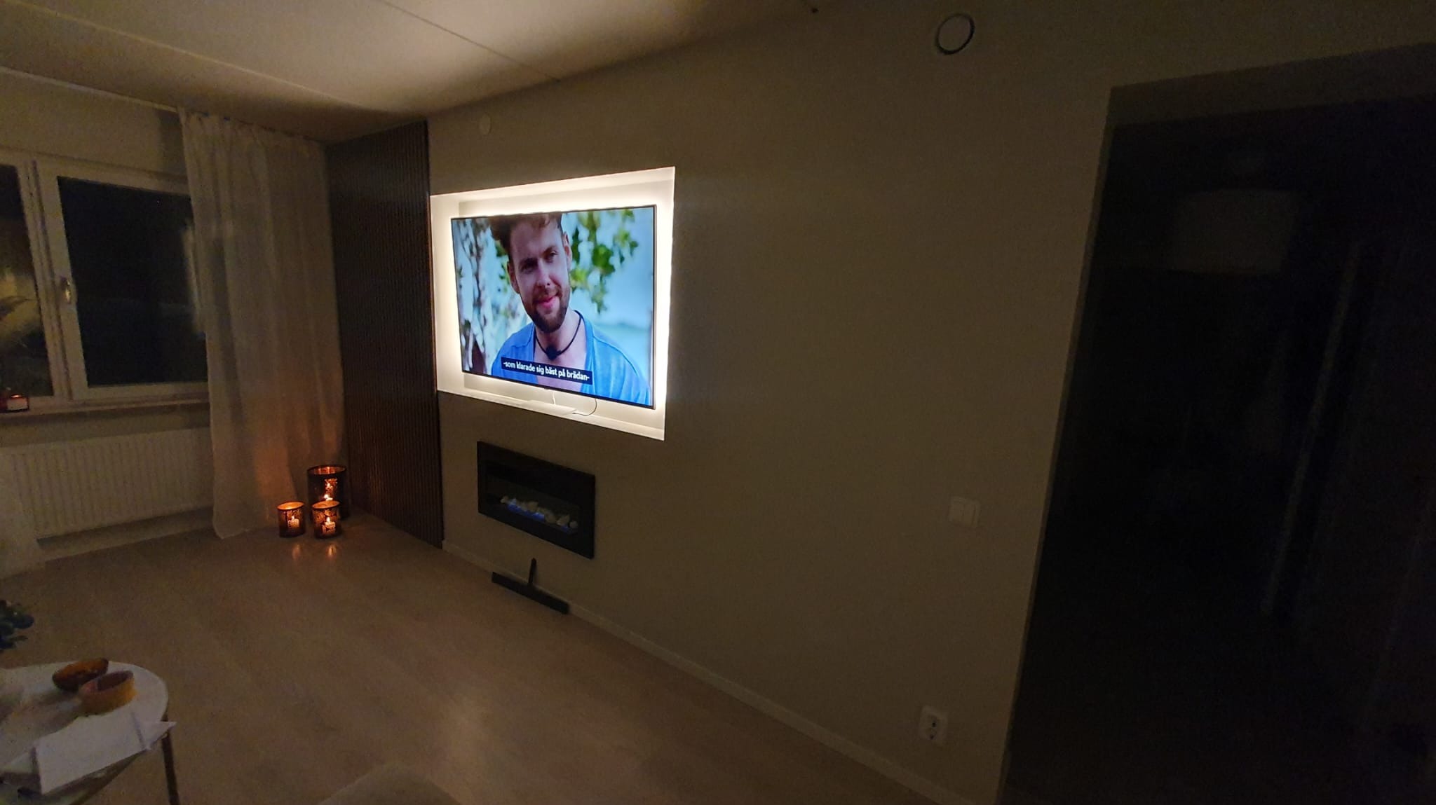 Telewizor z podświetleniem LED zamontowany na ścianie nad elektrycznym kominkiem, widoczne dekoracyjne świece i fragment okna z zasłoną.