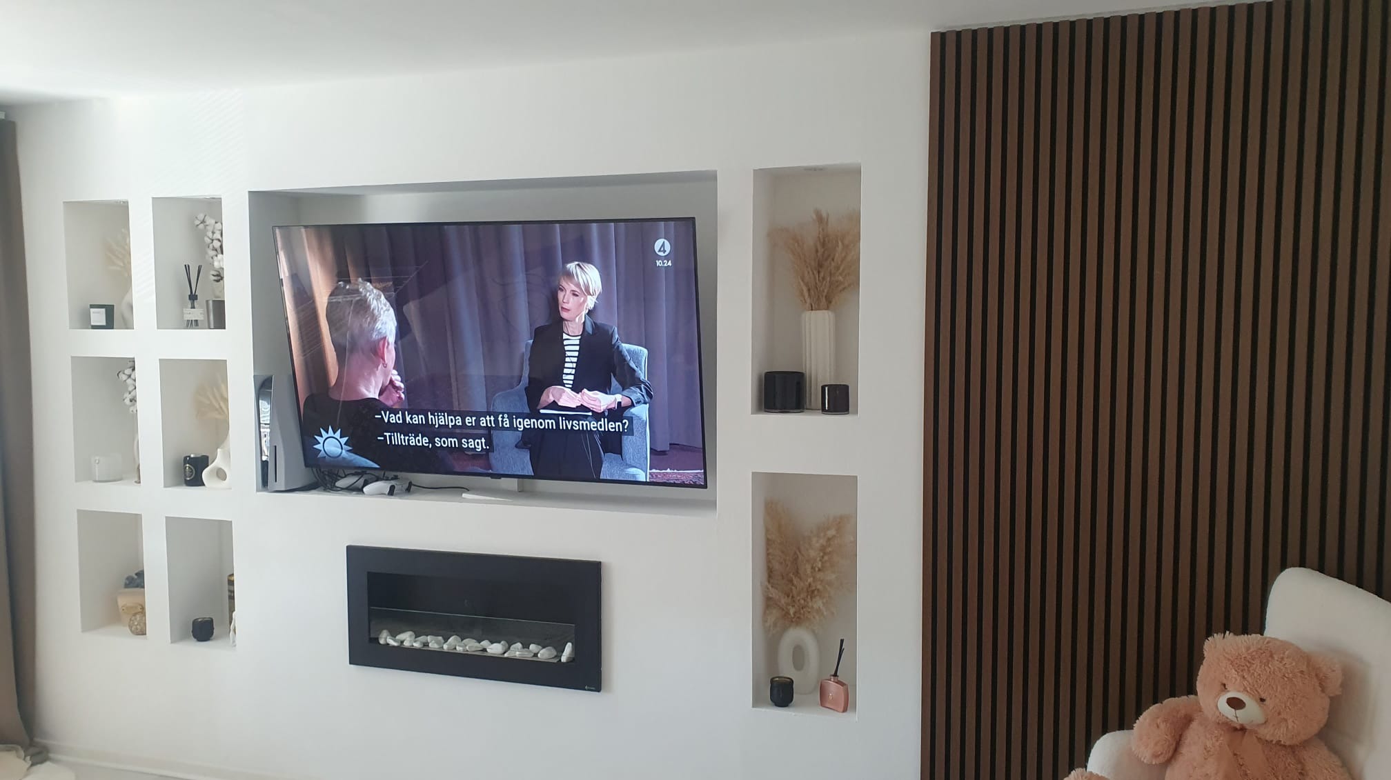 Telewizor wbudowany w ścianę z wnękami dekoracyjnymi, obok pionowy panel z drewnianych listew. W dolnej części czarny, prostokątny kominek elektryczny z białymi kamieniami.