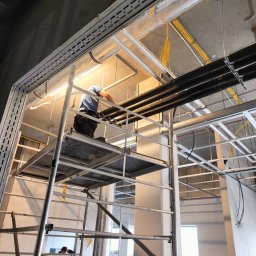 JAPKO MAŁGORZATA GRÜNING - Pracownik na rusztowaniu montuje rury instalacji chłodzenia w surowym wnętrzu przychodni, widoczne przewody elektryczne i elementy konstrukcyjne pod sufitem.