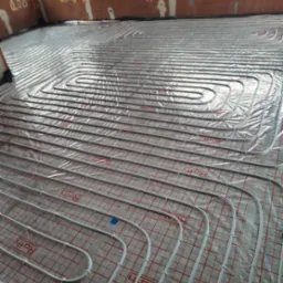 Białe rury ogrzewania podłogowego marki Roth ułożone w pętle na folii z czerwoną siatką, widoczne fragmenty muru z cegły dziurawki i okna.