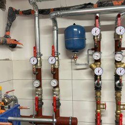 JAPKO MAŁGORZATA GRÜNING - Złożona instalacja hydrauliczna z rurami, zaworami i manometrami na tle białych płytek. Widoczny zbiornik wyrównawczy i fragment instalacji w izolacji.