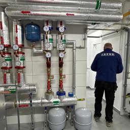 JAPKO MAŁGORZATA GRÜNING - Złożona instalacja grzewcza z rurami w izolacji, manometrami i zbiornikami wyrównawczymi. Widoczny hydraulik w uniformie z logo firmy. Szare płytki podłogowe.