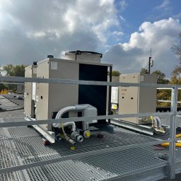 JAPKO MAŁGORZATA GRÜNING - Dwa klimatyzatory przemysłowe na dachu budynku, widoczne rury i elementy instalacji, kratownica zabezpieczająca. Błękitne niebo z chmurami w tle.