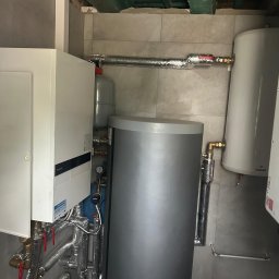 JAPKO MAŁGORZATA GRÜNING - Zestaw urządzeń grzewczych w kotłowni: piec, zbiornik buforowy, naczynie przeponowe. Rury w izolacji, szara płytka na ścianach, surowy strop. Nowoczesna instalacja CO.