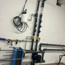 JAPKO MAŁGORZATA GRÜNING - Złożona instalacja hydrauliczna z czarnymi rurami, manometrami, zbiornikiem wyrównawczym i niebieskim wymiennikiem ciepła na tle białej ściany. Widoczne liczne złączki i zawory.