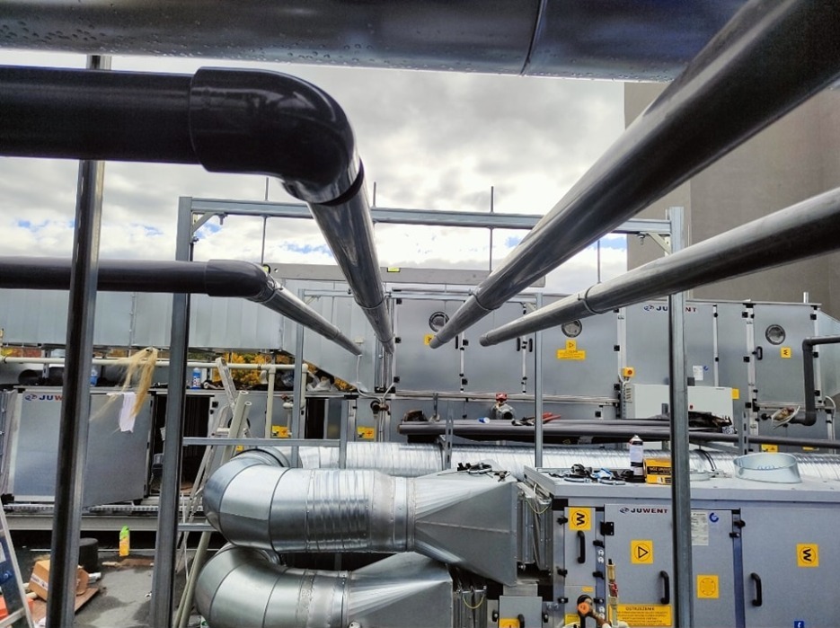 Instalacja przemysłowa wody lodowej w szpitalu, widoczne rurociągi z czarnego i srebrnego tworzywa oraz modułowe jednostki klimatyzacyjne Juwent z żółtymi tabliczkami ostrzegawczymi.