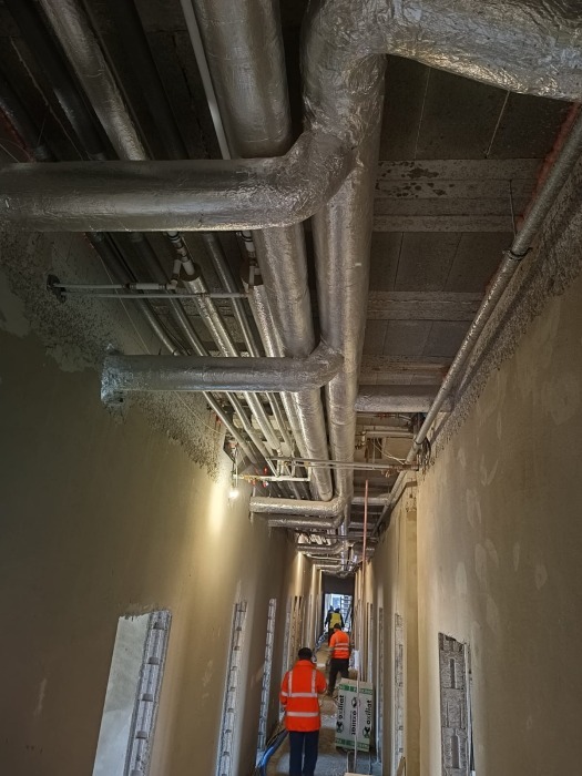 Korytarz w trakcie instalacji systemów CO, CWU, ZW, Gaz i PPOŻ, widoczne rury pod sufitem z izolacją, pracownicy w pomarańczowych kamizelkach i materiały budowlane.