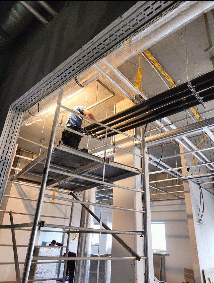 Pracownik na rusztowaniu montuje rury instalacji chłodzenia w surowym wnętrzu przychodni, widoczne przewody elektryczne i elementy konstrukcyjne pod sufitem.