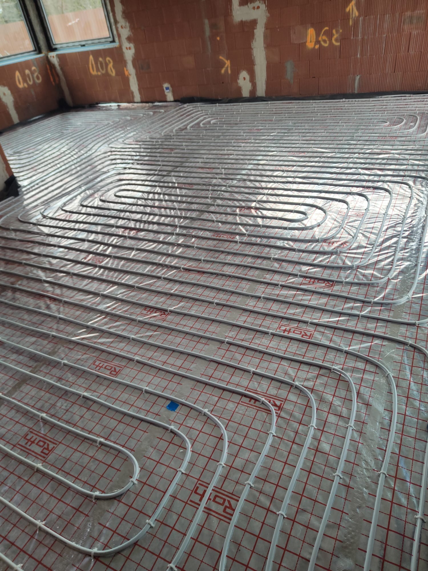 Białe rury ogrzewania podłogowego marki Roth ułożone w pętle na folii z czerwoną siatką, widoczne fragmenty muru z cegły dziurawki i okna.