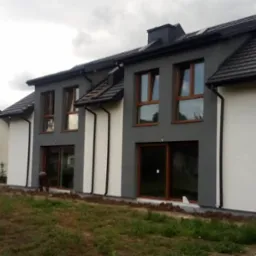 Dwukondygnacyjny dom z elewacją w odcieniach szarości i bieli, z brązowymi ramami okiennymi. Widoczne prace ziemne wokół budynku.