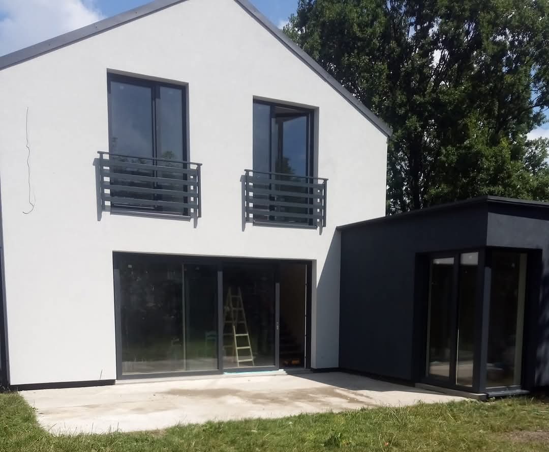 Nowoczesny dom z białą elewacją i grafitowymi oknami, widoczne balkony z barierkami, trawa przed budynkiem, drabina wewnątrz.