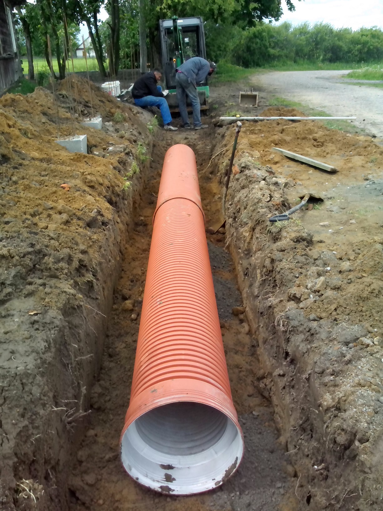 Pomarańczowa rura kanalizacyjna w wykopie w Otwocku, widoczna minikoparka i pracownicy w tle.