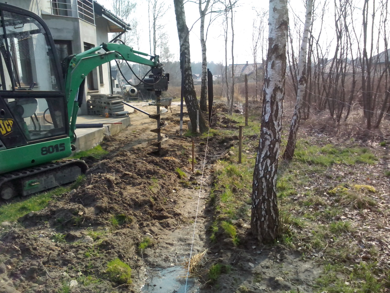 Zielona mini koparka z wiertnicą wierci otwory w ziemi pod słupki ogrodzeniowe, wytyczone sznurkiem, na tle brzozowego zagajnika i domu w budowie.