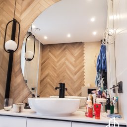 Finezja Wnętrz - Nowoczesna łazienka z umywalką nablatową, dużym okrągłym lustrem i ścianą wykończoną płytkami imitującymi drewniany parkiet ułożony w jodełkę; widoczne wiszące lampy z okrągłymi kloszami i logo...