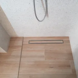 Nowoczesny odpływ liniowy w kabinie prysznicowej z beżowymi płytkami imitującymi drewno na podłodze i ścianami wykończonymi jasnym lastryko, widoczny fragment węża prysznicowego.
